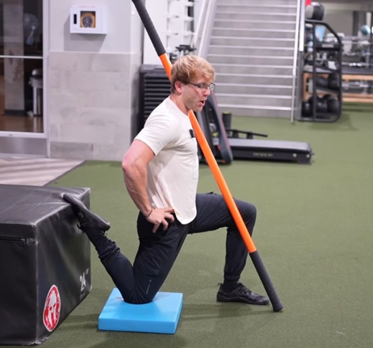Athlete in a split-stance stretch, rear foot on a box, holding an orange dowel
